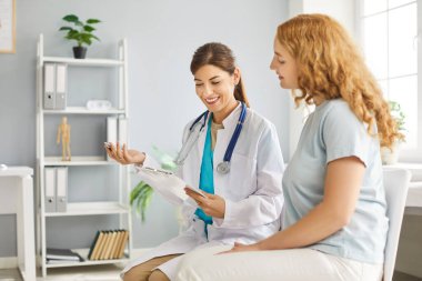 Elinde randevu ile rapor tutan genç doktor hasta bir kadın hastaya reçete yazıp muayene sırasında klinikte oturuyor. İlaç ve sağlık hizmetleri kavramı.