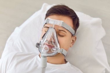 Hastane koğuşunda oksijen maskesiyle yatan ve oksijen tedavisi gören genç, baygın bir adamın fotoğrafını çek. Aspiratörlü erkek hasta komada yatıyor. Durumu kritik..
