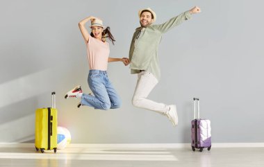 Komik, heyecanlı şapkalı mutlu çift tatil gezisine hazırlanıyor. Neşeli gülümseyen adam ve kadın turistler gri duvar arkasında bavullarıyla zıplıyorlar. Tatil ve yaz yolculuğu konsepti.
