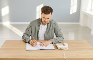 Genç adam iş ararken meşgul, sabit bir telefonla arama yapıyor ve not defterine bilgi yazıyor, boş bir odada masa başında oturuyor, ciddi bir ifadeye sahip, iş bulmaya çalışıyor.
