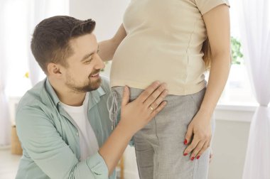 Evdeki oturma odasında hamile kadınların karnına bakıp gülümseyen genç adamın portresi. Karısının hamileliğinin tadını çıkaran sevgi dolu bir koca. Bebek bekleyen bir çift..