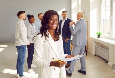 Mutlu bir gülümseyen Afro-Amerikan iş kadını liderinin portresi gururla arka planda şirket çalışanlarından oluşan bir ekiple kameraya bakıyor..