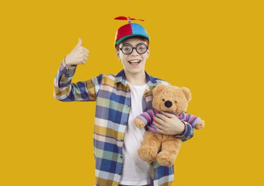 Küçük çocuk sarı, mavi, beyaz ekose gömlek, komik kırmızı pervane bere ve yuvarlak gözlük turuncu arka planda izole edilmiş duruyor, oyuncak ayıyı tutuyor, başparmak işareti yapıyor ve gülümsüyor.