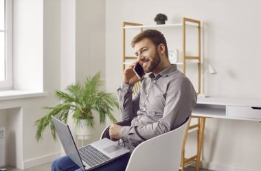 Dizüstü bilgisayarı olan, cep telefonuyla konuşan, koltukta oturan ve modern bilgisayar kullanan neşeli, güler yüzlü, sakallı bir iş adamı. Evde çalışmak, serbest çalışma kavramı.