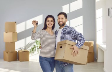 Yeni evin anahtarlarını gösteren aile çifti, kameraya gülümsüyor. Genç ve mutlu eşler oturma odasında arkalarında karton kutularla poz veriyorlar. Taşınma günü, emlak konsepti.