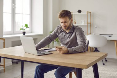 Bilgisayarlı, cep telefonuyla çalışan, modern ofisteki çalışma masasında oturan, kendine güvenen sakallı genç bir iş adamının portresi. Akıllı telefonu olan konsantre bir Ompany çalışanı..
