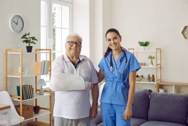 Mutlu yaralı erkek hasta portresi ve kadın doktor. Kolunda askı olan yaşlı bir adam ve mavi üniformalı genç bir kadın cerrah bir arada durup gülümsüyor. Yaralanma tedavisi kavramı