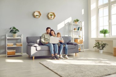 Genç ve mutlu bir ailenin modern bir dizüstü bilgisayar kullanarak koltukta oturan küçük bir kız çocuğuyla tam boy fotoğrafı. Ebeveynler kanepede dinleniyor. İnternetten komik çizgi filmler izliyor ya da video konuşmaları yapıyorlar..