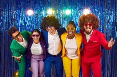 Neşeli, neşeli, parlak kostümler giymiş, şehvetli kıvırcık peruklar takmış, renkli büyük bardaklar takmış, dans eden ve parlak mavi arka planda oynaşan bir grup genç. Karnaval konsepti.