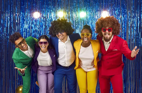 Neşeli, neşeli, parlak kostümler giymiş, şehvetli kıvırcık peruklar takmış, renkli büyük bardaklar takmış, dans eden ve parlak mavi arka planda oynaşan bir grup genç. Karnaval konsepti.