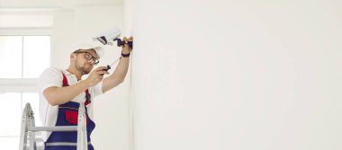 Erkek teknisyen ofisteki ya da evdeki beyaz duvara modern CCTV kameralarını yerleştirir ya da tamir eder. Merdivende tornavidalı bir adam akıllı CCTV izleme sistemini ayarlıyor. Uzayı kopyala Web pankartı.