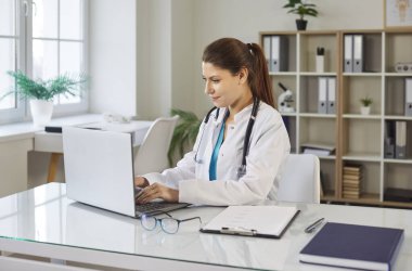 Beyaz üniformalı, gülümseyen, dost canlısı bir bayan doktor masada oturuyor ve tıp ofisinde dizüstü bilgisayarla çalışıyor. Steteskop takan neşeli kadın doktor klinikte çalışıyor..