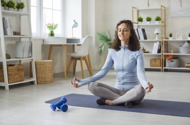 Yoga yapan rahat bir genç kadının portresi lotus pozuyla yerde oturuyor. Gözleri kapalı beyaz bir kız oturma odasındaki spor minderinin üzerinde meditasyon yapıyor. Sağlıklı yaşam tarzı.