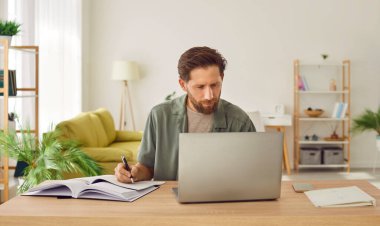 Kendine güvenen sakallı genç iş adamının portresi iş yerindeki masasında laptopuyla çalışıyor ya da not defterinde bilgisayar monitörüne bakarak notlar alıyor.