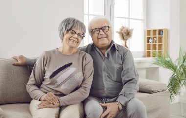 Evdeki oturma odasındaki kanepede oturan mutlu bir son sınıf aile çifti, karı koca portresi. İki gülümseyen gri saçlı emekli adam ve kadın emekliliğin tadını çıkarıyorlar..