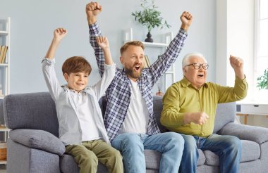 Üç nesil boyunca erkekler TV 'de heyecanlı spor müsabakaları izleyerek enerjik bir şekilde tezahürat yaptılar. Küçük çocuk, babası ve büyükbabasıyla tezahürat yapıyor. Kanepede oturuyor ve kutlama için ellerini kaldırıyor..