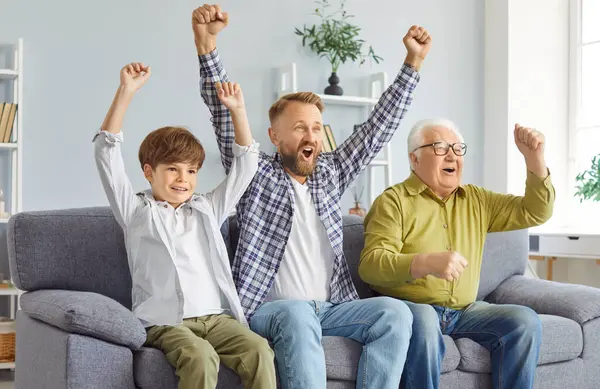 Üç nesil boyunca erkekler TV 'de heyecanlı spor müsabakaları izleyerek enerjik bir şekilde tezahürat yaptılar. Küçük çocuk, babası ve büyükbabasıyla tezahürat yapıyor. Kanepede oturuyor ve kutlama için ellerini kaldırıyor..