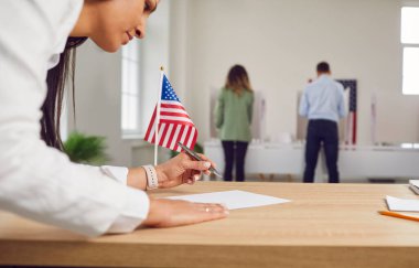 Oy verme merkezinde çalışan genç bir kadın. Amerikan seçmenlerinin oy verme kabininde arka arkaya oy kullanması ve oy verme kabininde ABD bayraklarının olması. Seçim günü ve demokrasi kavramı.