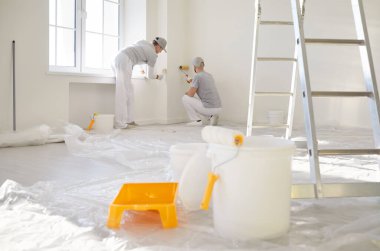 Boya silindiri, kova, tepsi ve merdiven, arka planda çalışan iki boyacının yenileme sürecini sembolize ediyor. İnşaat çalışmaları sırasında duvarlara yeni boya uygulayan işçiler.