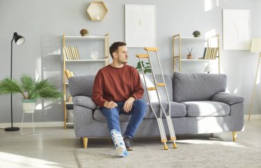 Koltuk değnekleriyle evde oturan üzgün genç adam rehabilitasyonda bacağı alçılı ve yaralı. Hasta kazadan sonra askı ya da bandaj takıyor. İyileşme ve rehabilitasyon konsepti.