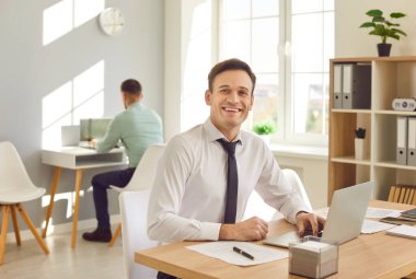 Ofiste dizüstü bilgisayarda yazan ve kameraya neşeli bakan gülümseyen genç iş adamı portresi. Çalışma masasında çalışan neşeli bir erkek şirket çalışanı..