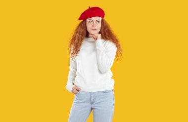 Kıvırcık kızıl saçlı, düşünceli, şaşkın, kırmızı bereli, beyaz kazaklı, sarı arka planda poz veren, şaşkın bir kız. Çenesine dokunuyor karar veriyor ya da sorunu düşünüyor...