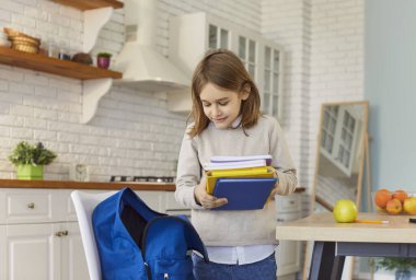 Küçük bir öğrenci okul öncesi ya da öğrenme aktivitelerini düzenlemek için sırt çantasına bir yığın kitap koyuyor, modern mutfak ortamında duruyor, küçük şirin öğrenci yeni bir çalışma gününe hazırlanıyor.