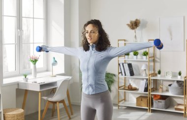 Küçük dambıllarla egzersiz yapan, güç antrenmanı sırasında kollarını yana kaldıran odaklanmış genç bir kadın. Spor giyim sektöründe atletik kadın spor yapıyor ve spor yaparken vücut kaslarını geliştiriyor..