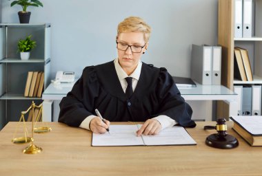 Profesyonel kıyafetli ciddi bir kadın hakim. Siyah cüppeli. Mahkeme masasındaki evrakları gözden geçiriyor. İnceleme, hukuki ve hukuki ayarların tartışılması, avukat danışmanlığı, dava hazırlığı.
