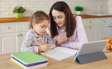 Anne ve kız tablet bilgisayarla internet üzerinden öğrenme faaliyetlerini öğreniyorlar, birlikte yemek, mutfak veya yemek salonunda oturuyorlar, ev ödevlerine odaklanıyorlar, küçük bir anaokulu kızına yardım eden bayan öğretmen. 