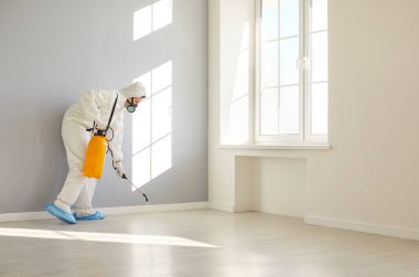 Modern boş odada böcek ilacı sıkan, dezenfekte eden, dezenfektan spreyiyle böcek ilaçlayan, temiz bir ortam sağlayan, beyaz iş kıyafetlerinde uzman bir ilaçlamacı.