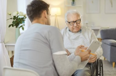 Kameraya bakan erkek hemşire, elinde tablet, tekerlekli sandalyedeki engelli yaşlı adama analiz sonuçlarını gösteriyor, reçeteyi açıklıyor, rehabilitasyondayken ona bakıyor.