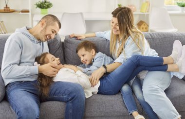 Genç ve mutlu bir aile oturma odasındaki büyük gri kanepede dinleniyor ve eğleniyor. Gülen ebeveynler ve şakacı oğul küçük kızları gıdıklıyor ve onlarla dalga geçiyor, hafta sonları evde eğleniyorlar..