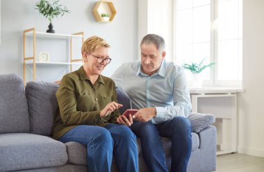 Evde oturma odasındaki kanepede dinlenen mutlu yaşlı çift birlikte boş zamanlarının tadını çıkarıyorlar. Online eğlence için bir cep telefonu kullanıyorlar, interneti keşfediyorlar, ve neşeli anları paylaşıyorlar..
