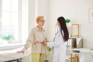 Klinikte düzenli tıbbi muayene sırasında kadın hastanın belini mezurayla ölçen genç kadın kardiyolog ya da laboratuvar önlüklü fizyoterapist. Kilo kaybı ve diyet konsepti.
