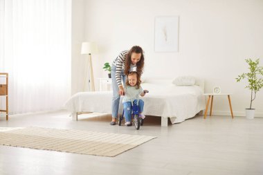 Genç, neşeli bir anne, mutlu, gülümseyen küçük kız çocuğunu evdeki oturma odasında bisiklete binmeyi öğretiyor. Bayan ebeveyn içeride kızıyla oynuyor. Mutlu aile ve çocukluk kavramı.
