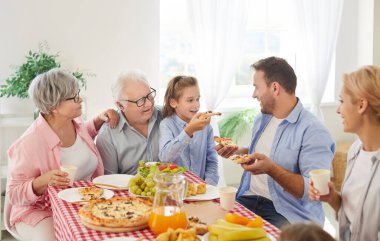 Mutlu bir aile hep birlikte yemek yiyor. Gülümseyen anne, baba, büyükbaba, büyükanne ve küçük bir çocuk evde bir masada oturuyor, yemek yiyor, pizza yiyor ve sohbet ediyor. 