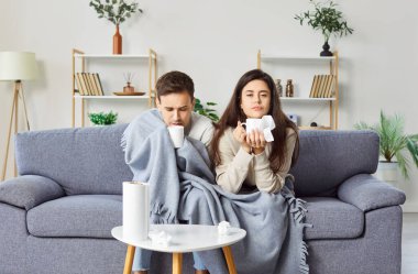 Hasta karı koca sıcak çay içip oturma odasındaki kanepede oturuyorlar. Ekoseyle kaplanmış, üşütmüş ve grip belirtileri gösteren genç, sağlıksız bir aile. Sağlık ve Tedavi.