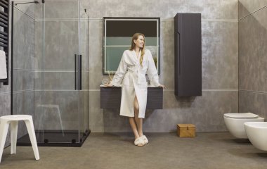 Beyaz bornoz giyen, modern banyoda dinlenen ve evde sabah keyfine varan neşeli ve çekici genç bir kadının tam boy portresi. Güzellik, sağlık ve hijyen konsepti.