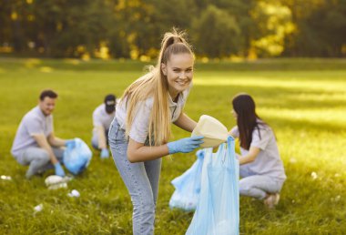 Kameraya bakan gülümseyen genç bir kadının portresi. Elinde çöp torbalarıyla gönüllü olarak dışarıdaki yaz parkındaki çimlerden çöp temizliyor. Çevre kirliliği ve ekoloji kavramı.