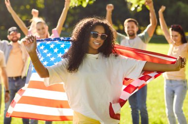 Amerika bayrağını tutan mutlu Afro-Amerikan kadın portresi Bağımsızlık Günü 'nü kutluyor yaz parkında arka planda gülen insanlarla birlikte duruyor. Vatanseverlik kavramı.