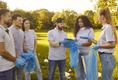 Bir grup gönüllü eldiven giyip, ellerinde çöp torbalarıyla yaz parkında çalışıyor ve dışarıda çöp topluyorlar. Doğadaki çevre kirliliğini temizleyen erkek ve kadınlar. Ekoloji kavramı.