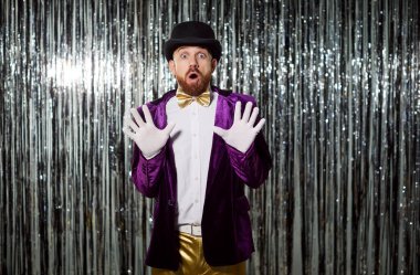 Komik müsrif adam, sihirbaz, parlak ceketli hokkabaz, şapkalı, başarıyı kutlamaktan heyecan duyuyor, esprili komik adam, metalik, parlak, uzun yağmurlu çelenk arka plan