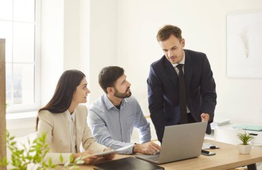 Parlak ofiste birlikte çalışan üç iş arkadaşı grubu. Masanın yanında duran takım elbiseli genç adam dikkatle dinleyen kadın ve erkeğe bir şey anlatıyor. Takım çalışması konsepti.
