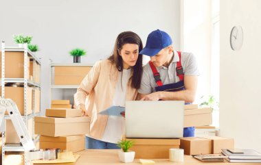 Depoda birlikte çalışan bir adam ve kadın, paket ve paketlerin envanterini çıkarmak için dizüstü bilgisayar kullanıyorlar. Takım çalışması, verimlilik ve iş operasyonları ve depolama alanında teknolojinin kullanımı.