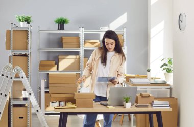 Depoda karton kutuların arasında çalışan kendine güvenen genç bir bayan çalışan. İşyerindeki stokları ve envanterleri kontrol eden bir kadın. Küçük işletme sahibi depoda müşteri için sipariş hazırlıyor.