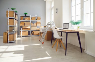Interior of empty work space with office desk in warehouse with shelves filled cardboard boxes of retail products. Business workplace used for shipping merchandise and stock inventory.