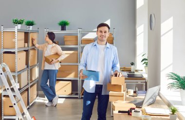 E ticaret işçileri. Çevrimiçi mağaza çalışanları depo odasında çalışıyor. Not panosu olan mutlu erkek envanter yöneticisi dizüstü bilgisayarı ve gönderilecek paket yığınıyla masanın yanında duruyor.