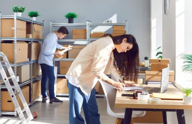 Çevrimiçi mağazada çalışan insanlar. Kadın ve erkek işçiler, küçük bir depoda, raflarda ve masalarda karton kutularla envanter kontrolü yapıyorlar. E ticaret KOBİ işletmesi