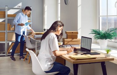 Bir erkek ve bir kadın online mağaza deposunda ya da postanede birlikte çalışıyor, envanter ya da işleme emirlerini yerine getiriyorlar. Çevrimiçi bir perakende veya posta ayarında depolama ve paketlerin yönetimi.